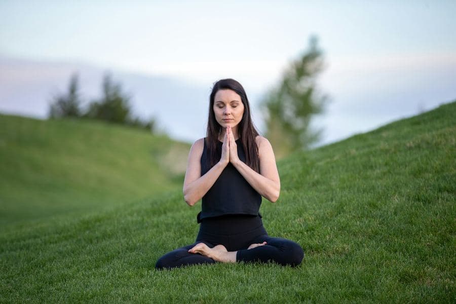 A woman sitting cross-legged on a green hillside, palms together, eyes closed